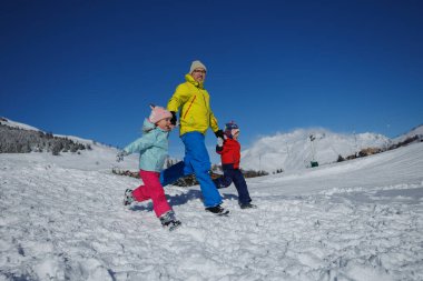 Üç kişi, baba ve çocuklar renkli kışlık ceketleriyle karlı bir yamaçta koşuyor, açık mavi gökyüzünün altında gevrek alp havasının tadını çıkarıyorlar.