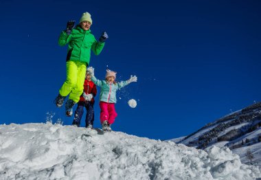 Bir grup çocuk, güneşli bir kış gününde, etrafları yemyeşil ağaçlarla çevrili, neşeyle karın içine atlarlar.