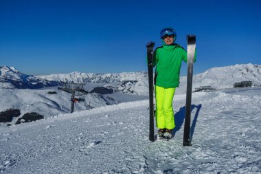 Bir kayakçı el değmemiş bir dağ yamacında dikey olarak yanında kayakları tutarak, uzaktan ışıldayan güneşli tepeleri seyreder.