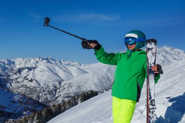 Bir kayakçı, bir omuz boyunca uzanan kayaklara yaslanır ve arkasında kar tozu dolu tepeler olan açık mavi gökyüzüne doğru selfie çubuğu uzatır.