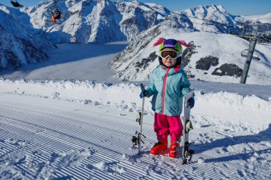 Pastel ceket giyen bir çocuk, rüzgarın süpürdüğü karlı sırtları ve açık gökyüzü olan kayakları sıkıca kavrıyor.