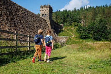 Sırt çantalı iki çocuk, büyük bir taşın yanında otlak bir patikada geziniyorlar. İngiltere 'nin Howden Barajı ve orman yamacı.