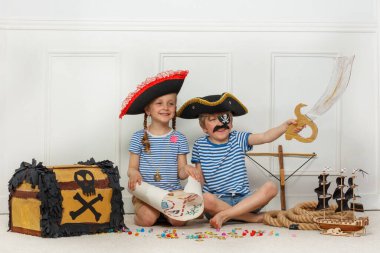 Yerde oturmuş korsan kıyafetli iki çocuk ellerinde bir hazine haritası, oyuncak bir kılıç, yanında da bir sandık ve bir model gemi tutuyorlar.