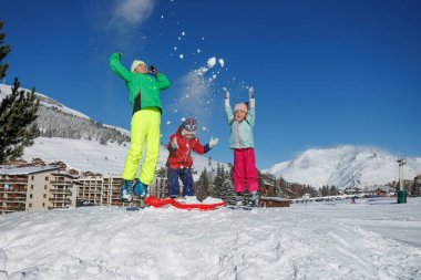 Bir genç ve iki çocuk karlı bir tepecikten atlayıp açık mavi gökyüzünün altında toz saçıyorlar.