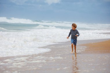 Genç bir çocuk kumlu bir sahilde arka planda köpüklü dalgalarla kıyı boyunca koşuyor.