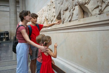Bir aile, bir müzede tarihi bir rahatlamayı gözlemlerken, anne ve çocuklar tatillerdeki sanat eserlerine işaret ediyorlar.