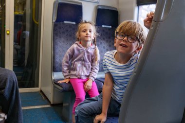 Trende oturan iki çocuk, mor kapüşonlu, pembe pantolonlu bir kız, gözlüklü ve çizgili gömlekli bir çocuk.