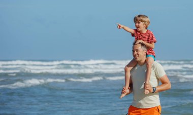 Neşeli bir çocuk, omuzlarında parlak mavi gökyüzü olan bir adamın sırtında gülüyor ve işaret ediyor. Okyanus dalgaları arka plan gibi.