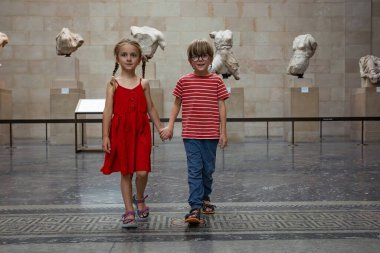 İki çocuk, kırmızı giyen kız ve çizgili giyen oğlan, el ele tutuşarak bir müzede dolaşıyorlar.