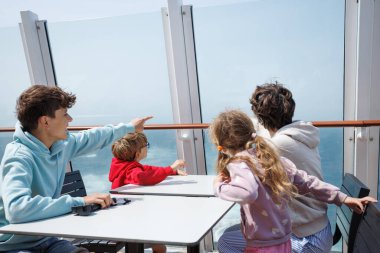 Bir feribot güvertesinde, bir turist ailesi bir masaya oturur ve Manş Denizi geçişi sırasında açık cam bariyerlerin arasından denizi izlerler.