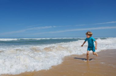 Genç bir çocuk kumlu sahil boyunca koşuyor dalgalar berrak mavi gökyüzünün altında okyanus kıyısında tatil yapmaktan mutlu bir şekilde çarpışıyor.