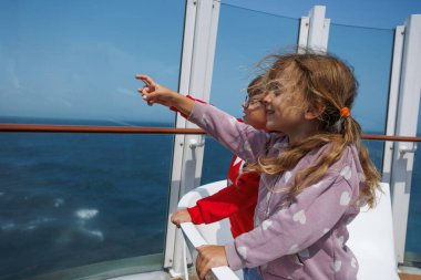Güneşli bir sahnede, arka planda engin bir okyanus manzaralı, güvertedeki bir masada oturan üç çocuk görülüyor.