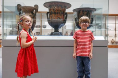 Bir çift çocuk, bir galeride çeşitli eserlerle çevrili cam bir ekranın içindeki etkileyici eski vazolara bakıyor.