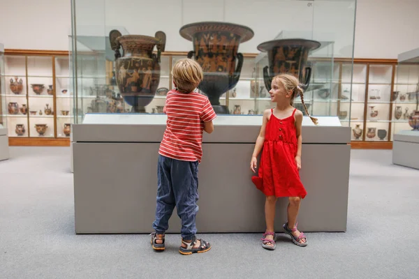 İki çocuk, bir müze sergisinde sergilenen büyük antik vazoları inceliyor. Gezinin karmaşık tasarımlarından büyülenmişler.