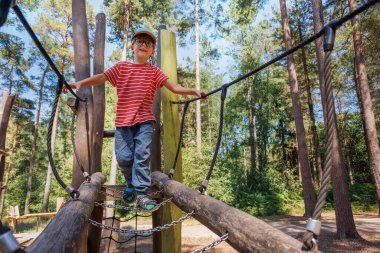 Küçük bir çocuk, uzun ağaçlar ve yeşilliklerle çevrili bir oyun alanındaki ahşap bir yapının üzerinde dengede durur. Yaz tatilinde arkadaşlarıyla açık havada güneşli bir günün tadını çıkarır.