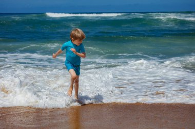 Genç bir çocuk güneşli bir sahilde neşeyle dalgalar arasında koşar, açık gökyüzü ve parlak güneşin altında hafif dalgalarla deniz kenarında neşeli bir günün tadını çıkarırken su sıçratır.