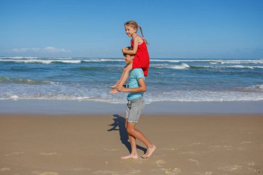 Bir erkek, tatillerde sahilde yürürken bir kızı sırtında taşır.