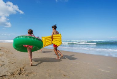 İki çocuk kumsalda neşeli bir şekilde koşarken açık mavi gökyüzünün altında dalgalar çarparken renkli şişme şamandıralar taşıyorlar.