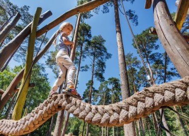 Genç bir kız, uzun ağaçlar ve açık mavi gökyüzüyle çevrili bir oyun parkındaki ip köprüde güven içinde dengede duruyor, maceracı ruhunu gösteriyor.