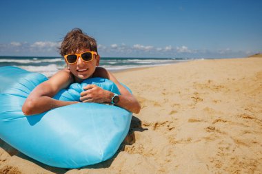 Genç bir çocuk kumsalda güneşli bir günün tadını çıkarırken etrafı kum ve okyanus dalgalarıyla çevrili, kabarık bulutlarla dolu parlak şişme bir şamandırada uzanır.