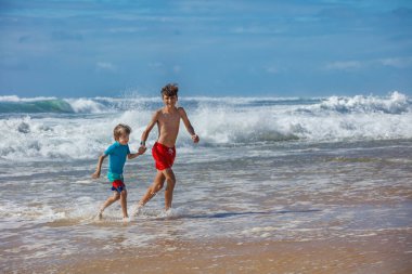 Yaz tatilinde kumsalda güneşli bir günün tadını çıkarırken, sahilde neşe içinde koşan iki çocuk, kıyı ve doğada eğlenceli, neşeli bir atmosfer yaratıyor.