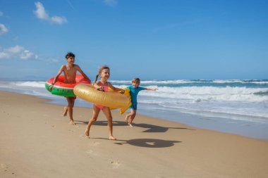 Üç çocuk kumsalda neşeyle koşuyor, ellerinde renkli şişirilebilir şamandıralar tutarken dalgalar arka planda berrak mavi bir gökyüzünün altına çarpıyor.