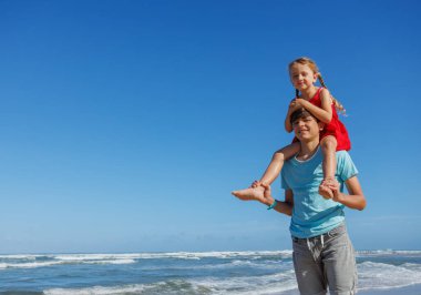 Genç bir kız, erkek kardeşinin omuzlarında eğlenceli bir anın tadını çıkarır. Kumsalda sırtına biner.
