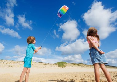İki çocuk kumsalda güneşli bir günün tadını çıkarırken renkli bir uçurtmayı açık mavi gökyüzü ve kabarık bulutlara doğru uçuruyorlar.