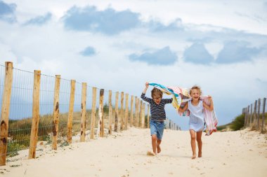 Yaz tatillerinde kumlu bir yolda neşeyle koşan iki çocuk, ellerinde renkli havlularla kumsalda güneşli bir günün tadını çıkarırken arka planda ahşap bir çit var.