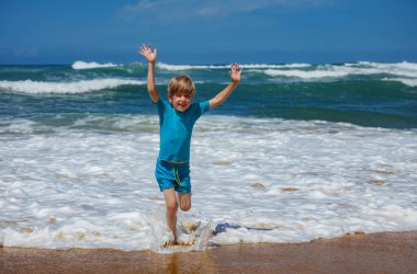 Genç bir çocuk sahilde güneşli bir günün tadını çıkarırken okyanus dalgalarında mutlu bir şekilde sıçrıyor. Deniz kenarındaki yaz etkinliklerinde eğlence ve heyecanın esansını yakalıyor.