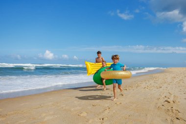 Üç çocuk kumsalda neşeli bir şekilde koşuyor, ellerinde renkli şişme şamandıralar, okyanus dalgalarının güneşli bir gününün tadını çıkarıyorlar.