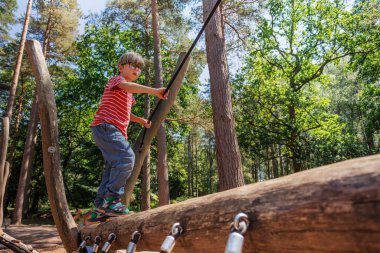 Küçük bir çocuk, ağaçlarla çevrili bir oyun parkındaki ahşap bir yapının üzerinde dengede duruyor. Yaz tatilinde fiziksel beceri ve güven geliştirirken doğada güneşli bir günün tadını çıkarıyor.