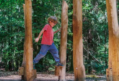 Bir çocuk, ağaçlarla dolu doğal bir ortamda çeviklik ve oyunculuk sergileyerek ağaçların arasındaki tahta direklerde ustalıkla denge kuruyor.