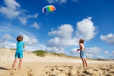İki çocuk kumsalda güneşli bir günün tadını çıkarırken mavi gökyüzünün arka planında renkli uçurtmalar uçuruyorlar, neşe ve neşe dolu bir atmosfer yaratıyorlar.