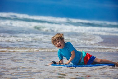 Genç bir çocuk sahilde sörf tahtasına uzanmış, dalgaların arka planda düşmesiyle neşeyle gülümsüyor, yaz tatilinde su aktiviteleri ve aile eğlencesi için mükemmel bir atmosfer yaratıyor.