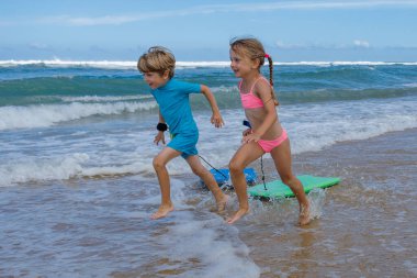 İki çocuk, erkek ve kız, güneşli bir sahil gününde ellerinde yeşil bir sörf tahtasıyla dalgalar arasında neşeyle koşuyorlar.