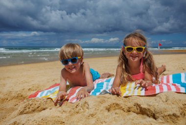 İki çocuk kumlu bir sahilde canlı havlular üzerinde uzanmış güneş gözlüğü takıyor ve bulutlu bir gökyüzünün altında, arka planda hafif dalgalar olan okyanusun tadını çıkarıyorlar.