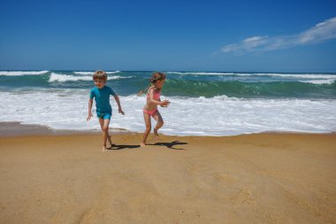 İki çocuk kumlu sahilde koşuyor, sıcak güneşin tadını çıkarıyorlar, dalgalar sıçrıyor, yazın eğlenceli ve kaygısız anlarını yaratıyorlar.