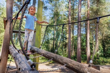 Genç bir kız güvenli bir şekilde bir orman oyun parkında ip köprüde yürüyor, yaz tatilinde doğada geçirdiği zamanın tadını çıkarıyor.
