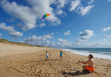 İki çocuk kumlu bir sahilde renkli bir uçurtma uçururken bir yetişkin onları mavi bir gökyüzünün altında seyrediyor.