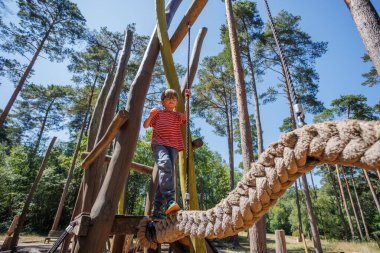 Bir çocuk, uzun ağaçlarla çevrili bir oyun parkında, ince bir iple ustaca dengede duruyor, açık havada güneş ışığı ve açık mavi gökyüzü ile doğal bir ortamda eğleniyor.