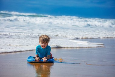 Genç bir çocuk kumsalda bir sörf tahtasına uzanmış, açık bir gökyüzünün altında, açık havada sörf yapmaya hazırlanırken gülümsüyor, dışarıda eğlenceli bir günün tadını çıkarıyor.