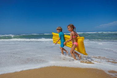 İki çocuk, kız sahilde koşar parlak sarı şişirilebilir bir şamandıra taşır etraflarını dalgalar kaplar açık mavi gökyüzü altında güneş ışığı ve neşe ile dolu bir yaz günü
