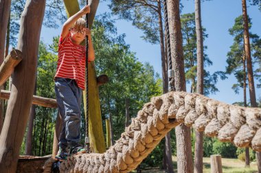 Genç bir çocuk güvenli bir şekilde bir orman oyun parkındaki ip köprüde duruyor, güneşli günün tadını çıkarıyor, uzun ağaçlar ve doğal manzarayla çevrili, keşfederken, eğlenirken.