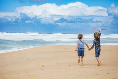 İki çocuk, kumlu bir sahil boyunca yürürler, biri kırmızı oyuncak uçak tutarken okyanus dalgaları arka planda, tüylü bulutlarla dolu parlak mavi bir gökyüzünün altında çarpar.