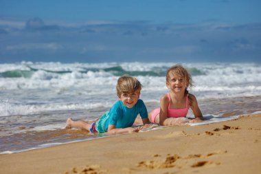 İki çocuk yaz tatilinde kumsalda güneşli bir günün tadını çıkarırken sığ sularda oynarken dalgalar, parlak ve berrak bulutlarla dolu bir gökyüzünün altında yavaşça kıyıya vuruyor.