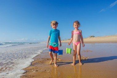 İki çocuk kumsalda ellerinde renkli kovalarla yürürken okyanus dalgaları ve kumlu sahilde güneşli bir günün tadını çıkarıyorlar.