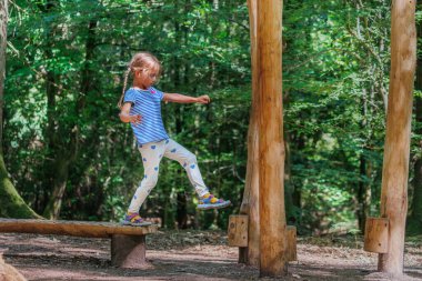 Genç bir kız, verimli yeşillikler ve uzun ağaçlarla çevrili bir orman bahçesindeki ahşap bir kirişin üzerinde ustalıkla denge kuruyor.