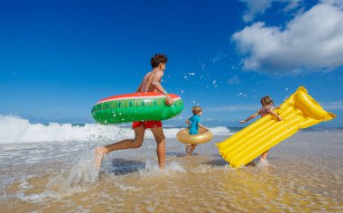 Üç çocuk arkadaş sevinçle plaj boyunca koşar, suya su sıçratır parlak mavi bir gökyüzünün altında kabarık bulutlarla renkli şişme şamandıralar taşırlar.