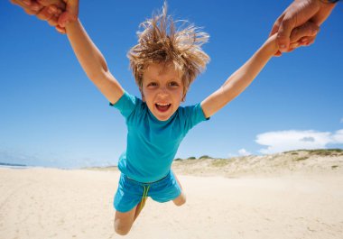 Saçı dağınık mutlu bir çocuk güneşli bir plajda oynarken ailesi tarafından sevinçle kaldırılır. Güzel bir açık hava ortamında aile eğlencesi ve mutluluğu tasasız bir anı yakalar.